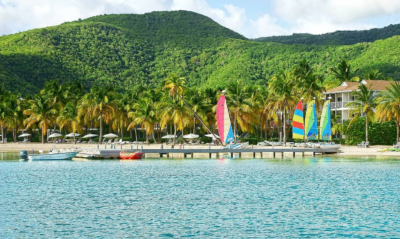 A view of the beach of the Carlisle Bay Resort. Beach umbrellas, sailboats, palm trees and a green hill behind.