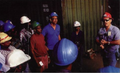 BSI Services Limited Trinidad, workmen in hard hats stand around another talking to them in a meeting.