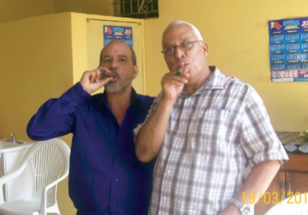 Two Caribbean Cable Company Limited workers posing for a photo, each holding a cigar to their mouth.