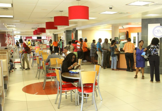 Sky Mall Barbados food court with people sitting at tables down the center eating food. With many more patrons standing in line in front of fast food restaurants to the right.