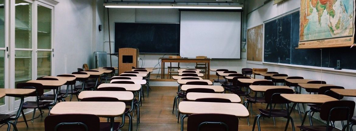 Rows of school desks with a podium and projection screen in the background. A map on the right over a chalk board.