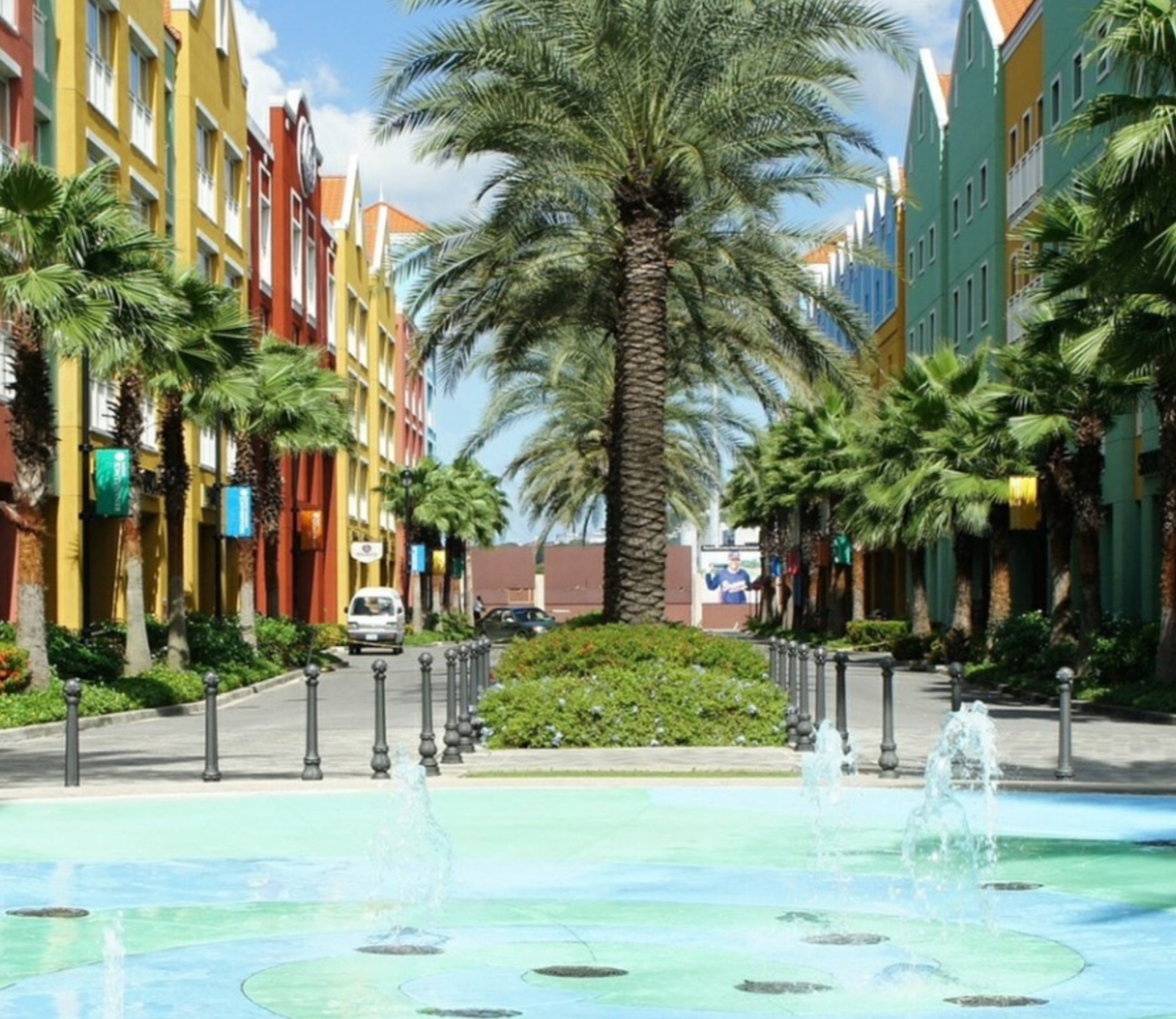 A beautiful street view with bright color buildings of blues, greens, oranges and reds lining both sides of a street with a median. Palms trees lining the street and in the foreground, a blue and cement swirl pattern on the ground with water fountains throughout. Property represented by Crye-Leike Real Estate Services.