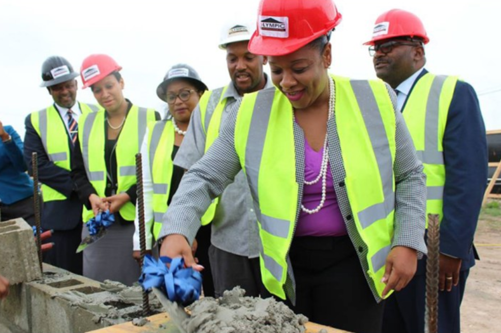 A group of people in hard hats and vests watching a woman in the foreground apply mortar to a row of bricks on a structure.