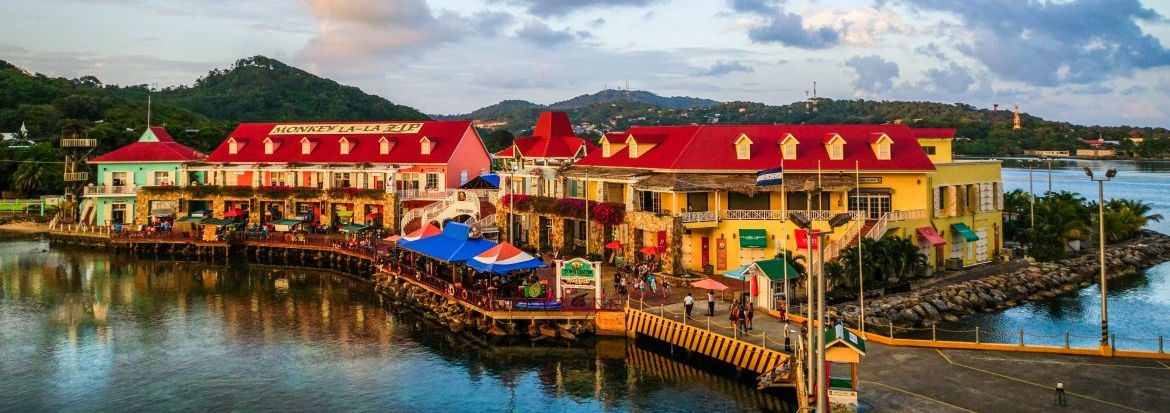 Roatan, a beautiful group of commercial buildings in the middle ground, located on the water with a dock in the foreground leading out from the businesses.