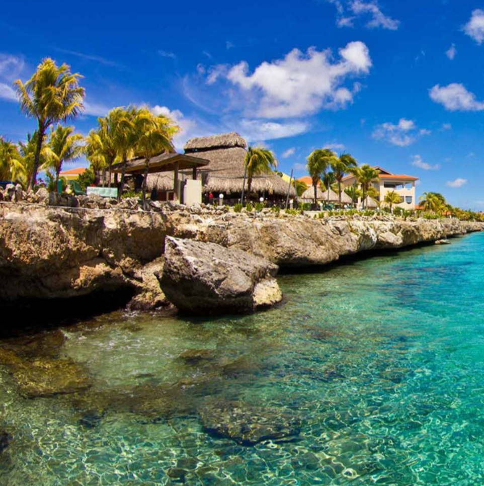 A beautiful blue sky with a few white fluffy clouds. Including waterfront buildings , one with a tiki style, located on the clear blue water and rocky shoreline.