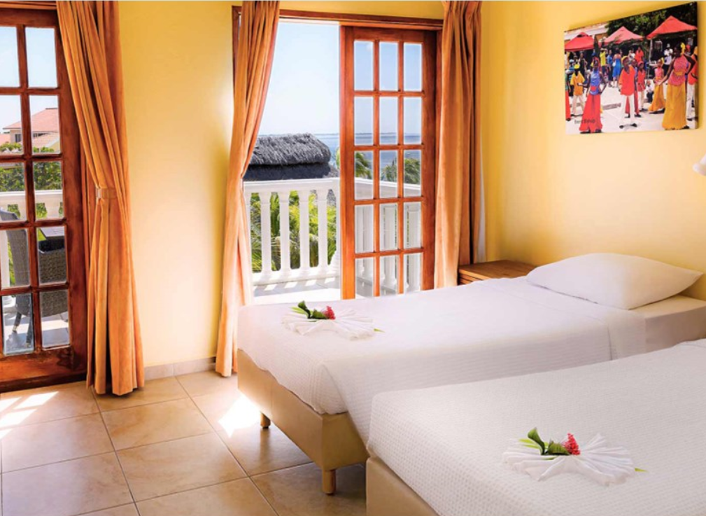 A well lit room with freshly made beds. Open balcony doors leading out to the Caribbean.