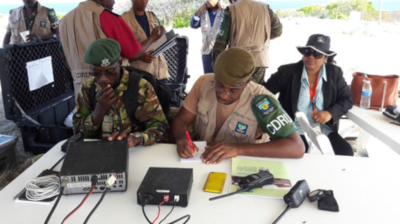 Men in military uniforms sitting at a table with communication devices on the table and people talking behind them.