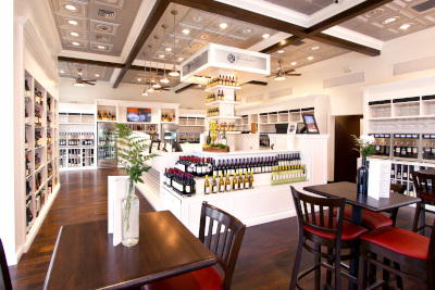 An example of work for Corporate Electric, showing a beautiful wine store with shelving around the outside of the room. Wood flooring with tables and chairs to sit in the foreground. In the middle is a square countertop showcasing more wine with a checkout area.