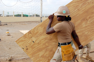 Incorporated Masterbuilders Association Jamaica. A woman in uniform with a hard hat and tool belt carrying a piece of plywood on a job site.