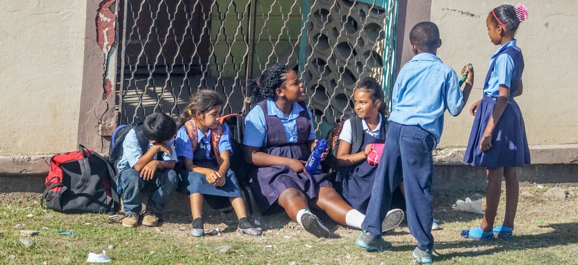 Dominica School children sitting and standing outside of a building on the grass together.