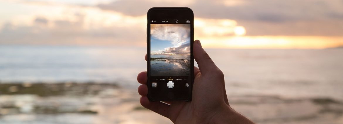 A hand holding a cell phone on a beach with a photo of the beach on the phone.