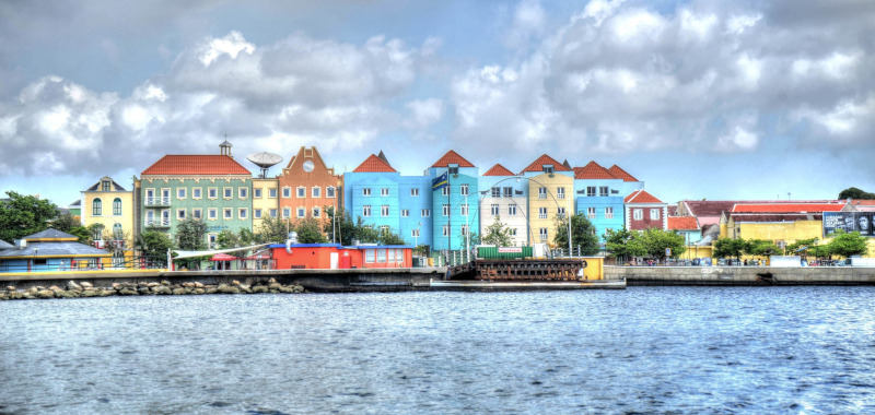 Willemstad ocean view of a row of colorful houses along the water with clouds above. Carribean Hotel &amp; Tourism Association.