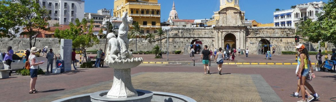 People walk around a fountain in Cartagena, with building behind.